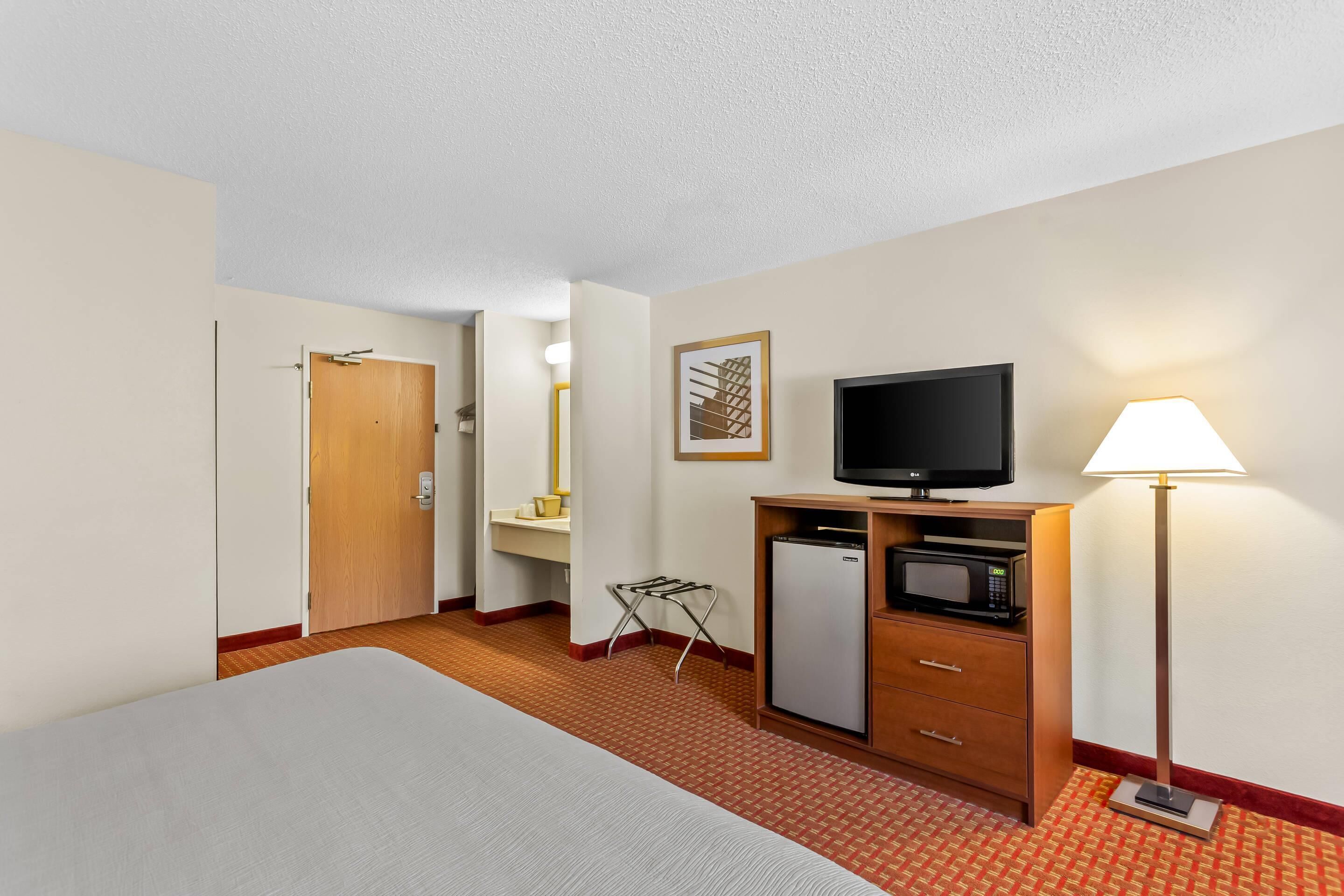 A hotel room with a bed, a television, a refrigerator, a lamp, and a small table.