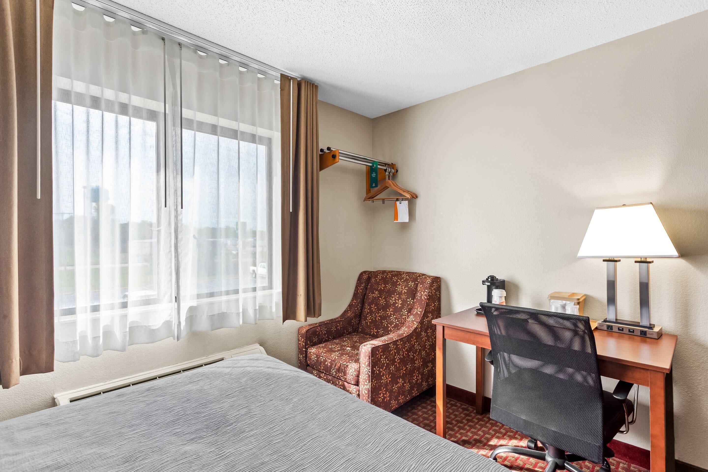 A hotel room with a bed, a desk with a lamp, a chair, and a window with curtains.