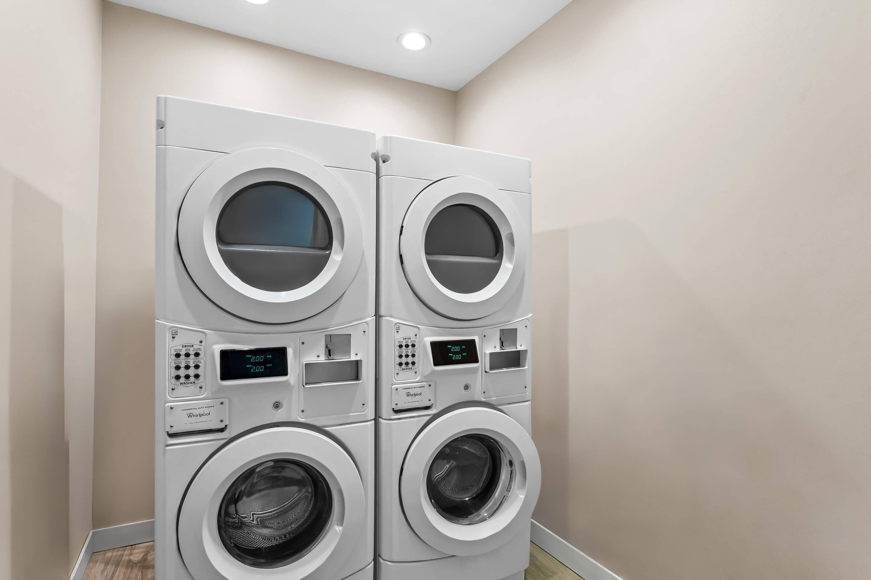 A four-door commercial laundry machine with a digital display, located in a room with a plain wall and a wooden floor.