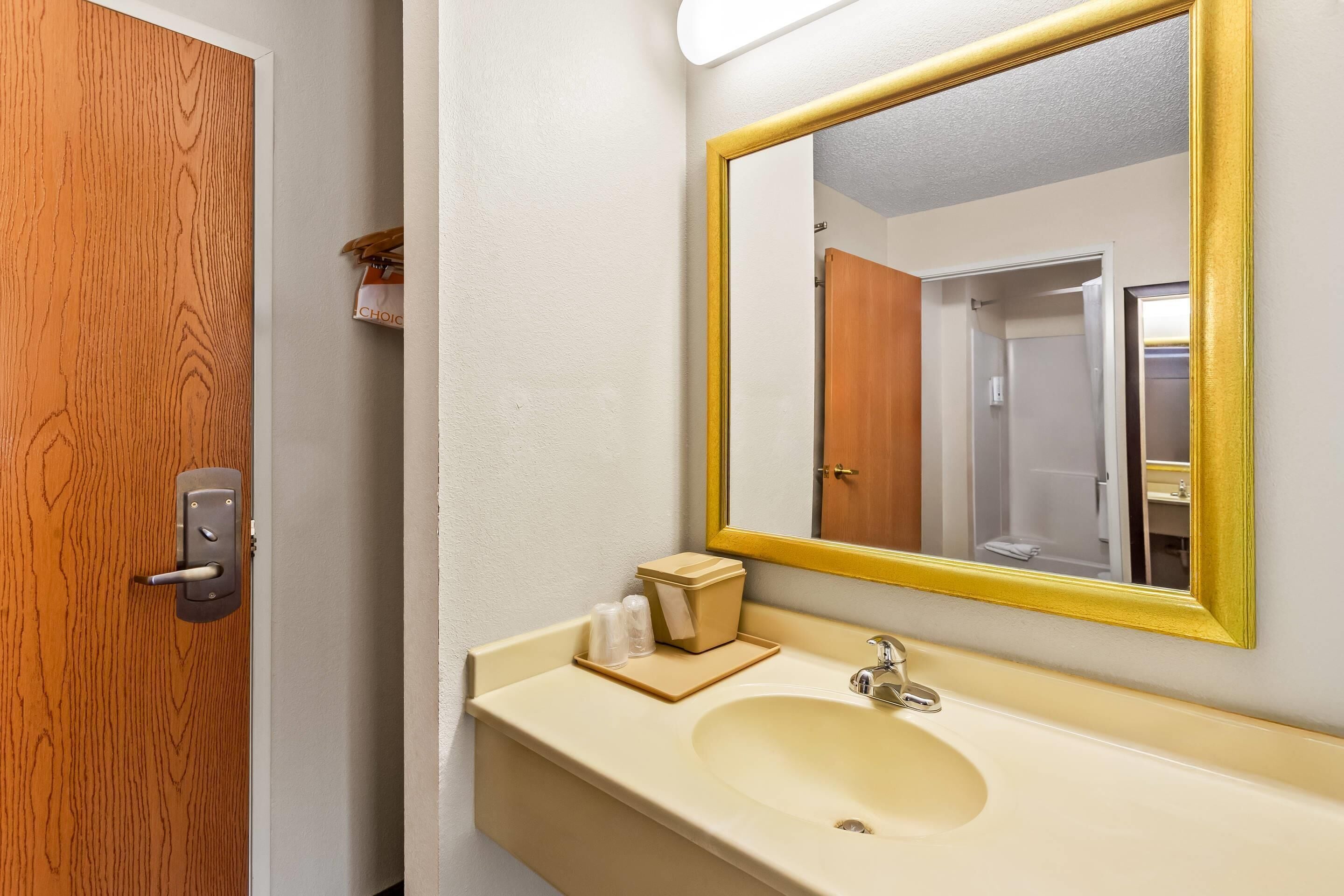 A bathroom with a wooden door, a beige sink, a mirror with a gold frame, and a towel rack.