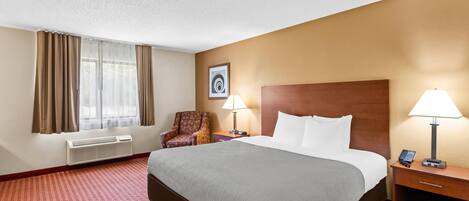 A hotel room with a large bed, two bedside tables with lamps, a chair, a wall-mounted air conditioner, and a framed picture on the wall.