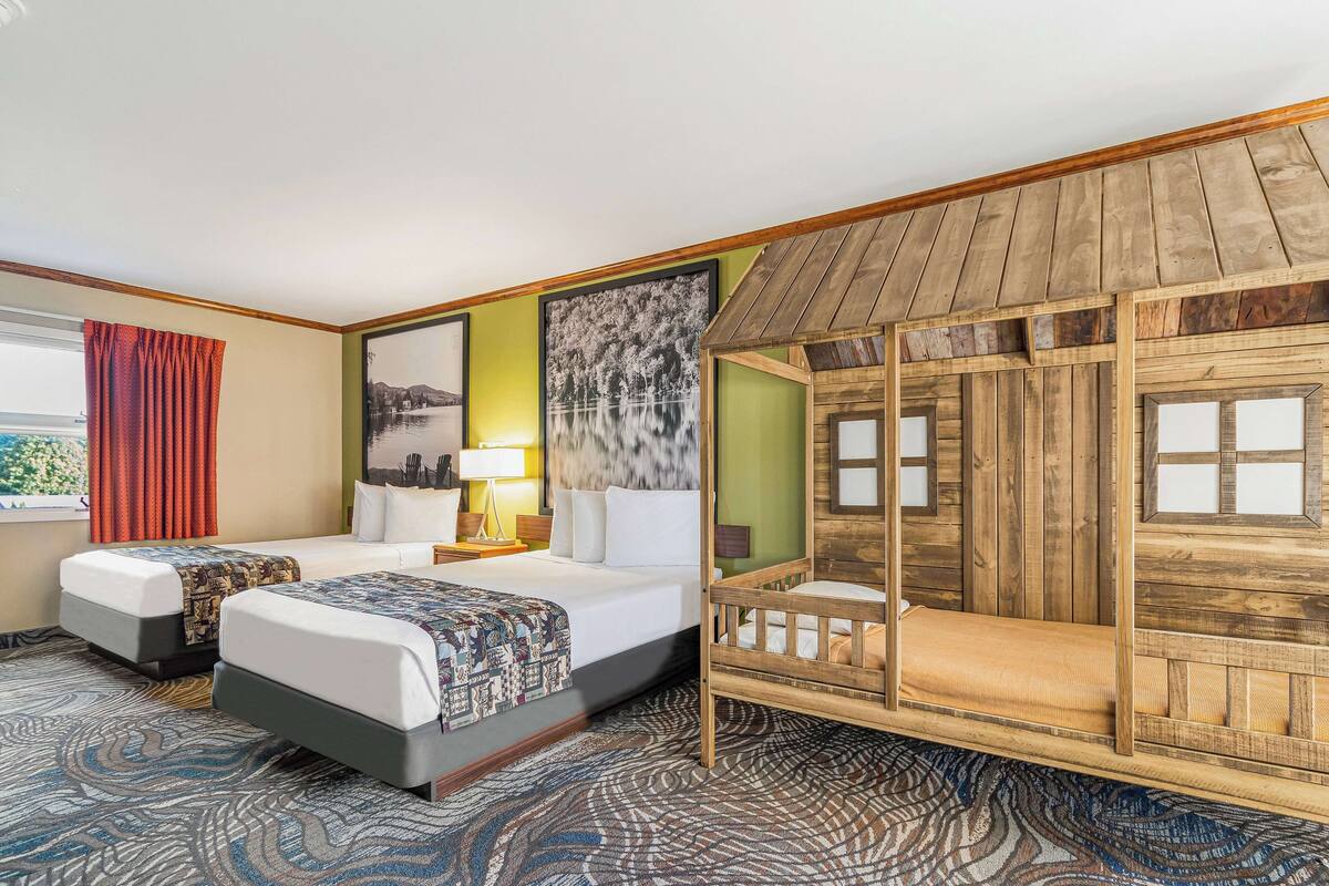 Une chambre d’hôtel avec deux lits, une maisonnette en bois, une grande fenêtre donnant sur un beau paysage et un tapis à motifs.