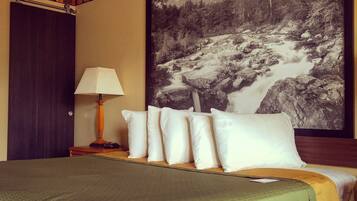 A hotel room with a bed, pillows, a lamp, a painting of a mountain stream, and a door.
