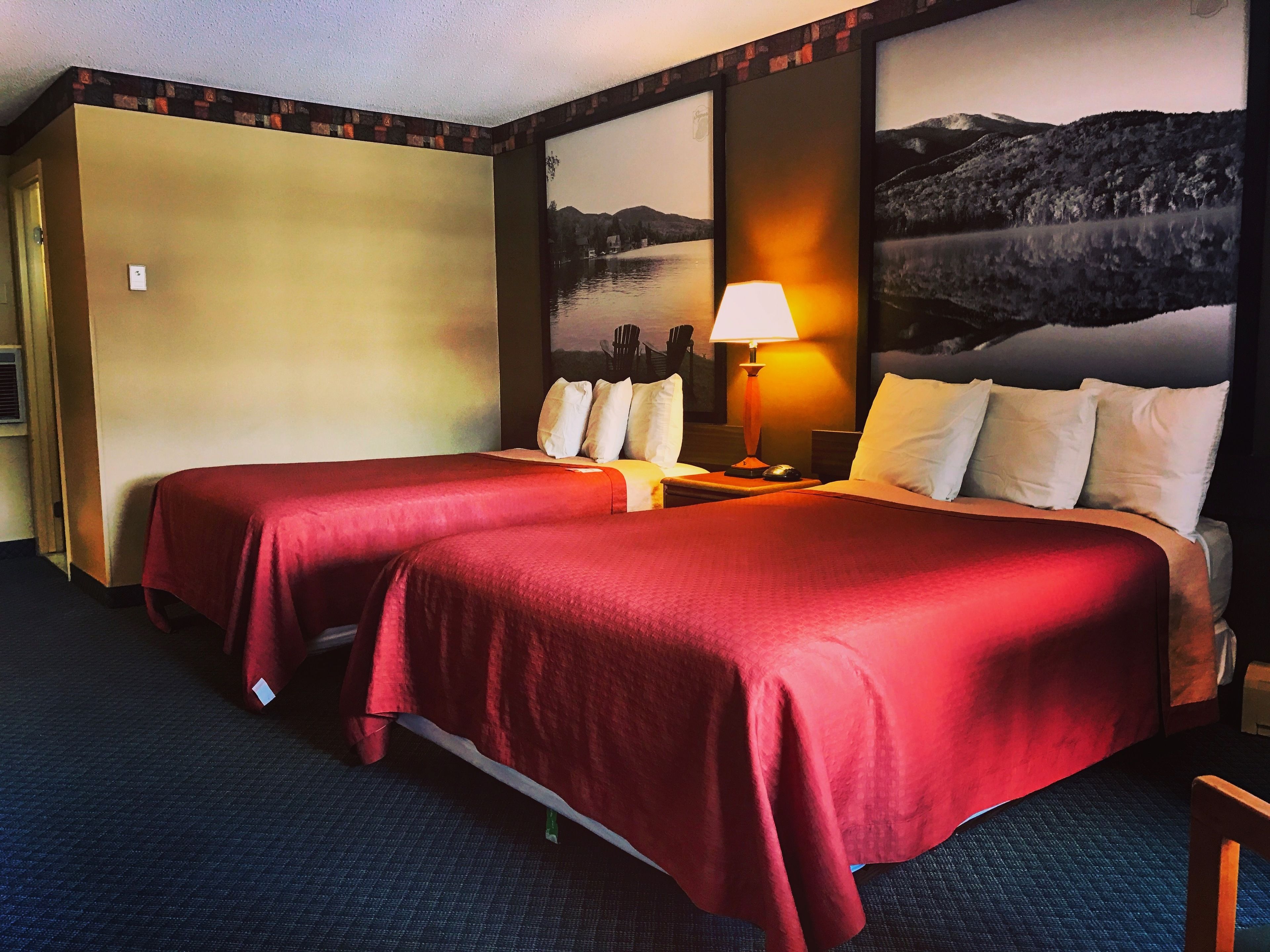 A hotel room with two beds, a nightstand, a lamp, and a painting of a mountain landscape.