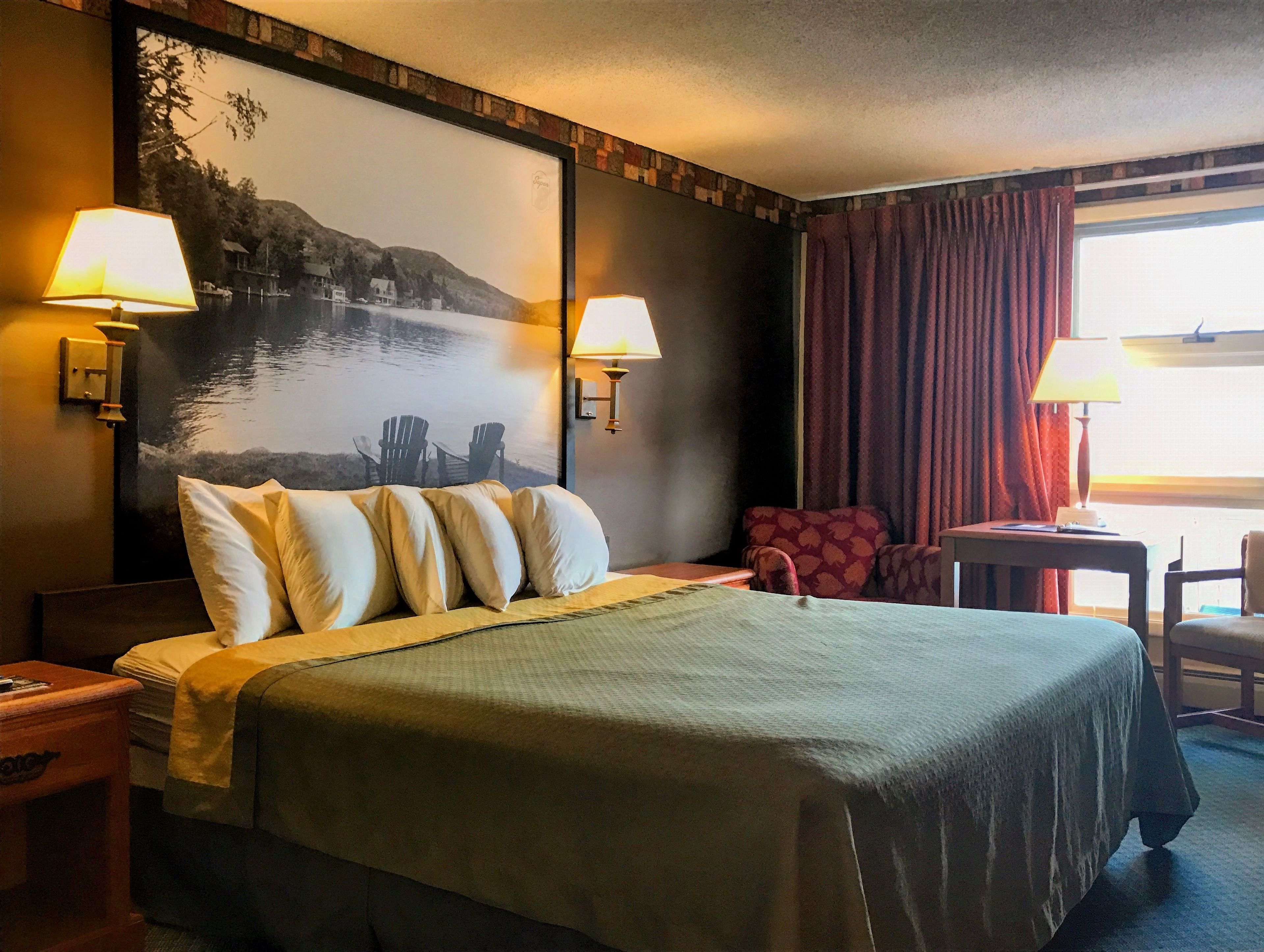 Camera d'albergo con un letto, due lampade a parete, una sedia, un tavolino e un'ampia finestra con tende.
