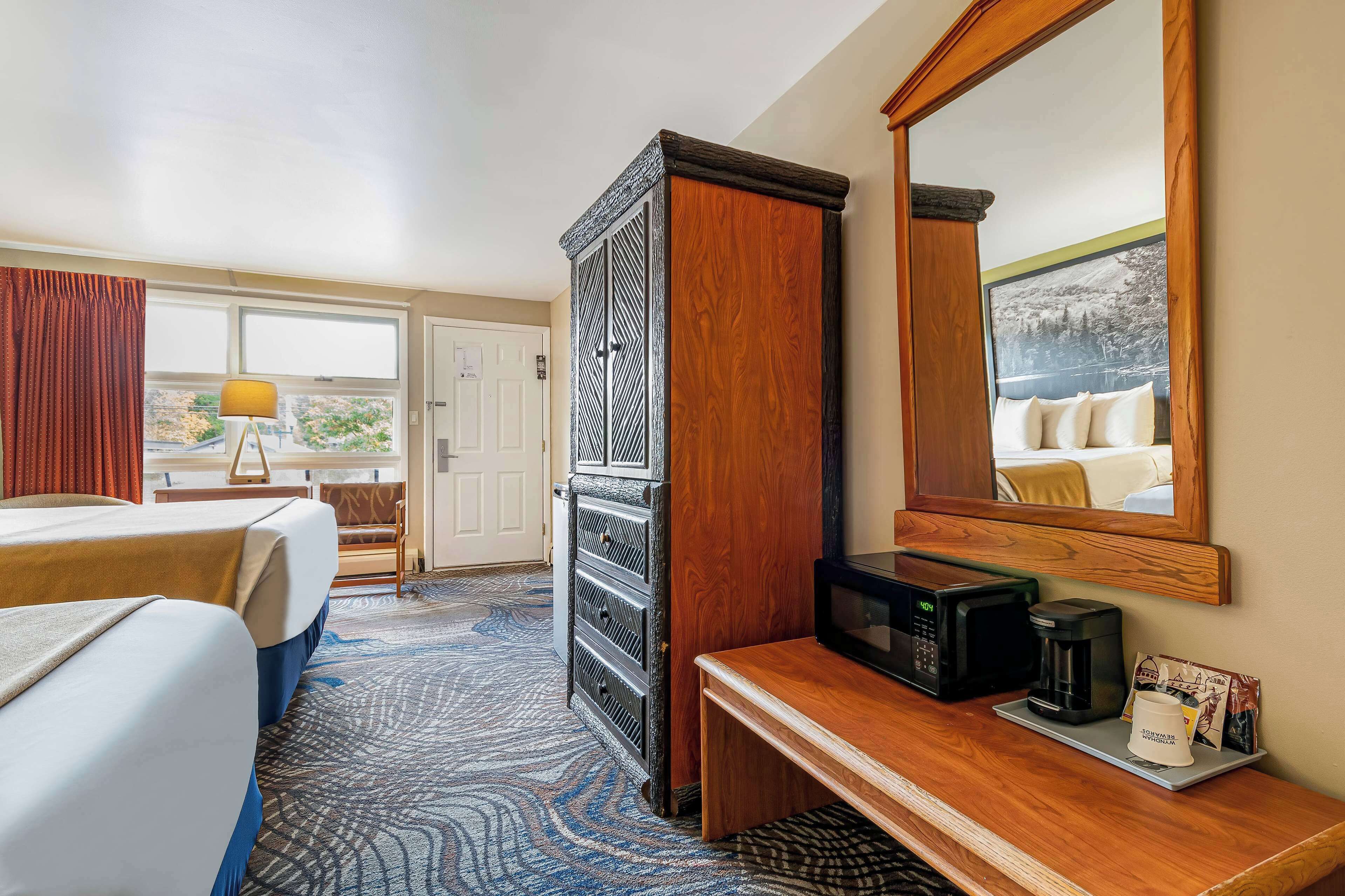A hotel room with two beds, a wooden cabinet, a coffee maker, a microwave, and a large mirror reflecting a bed and a window view.