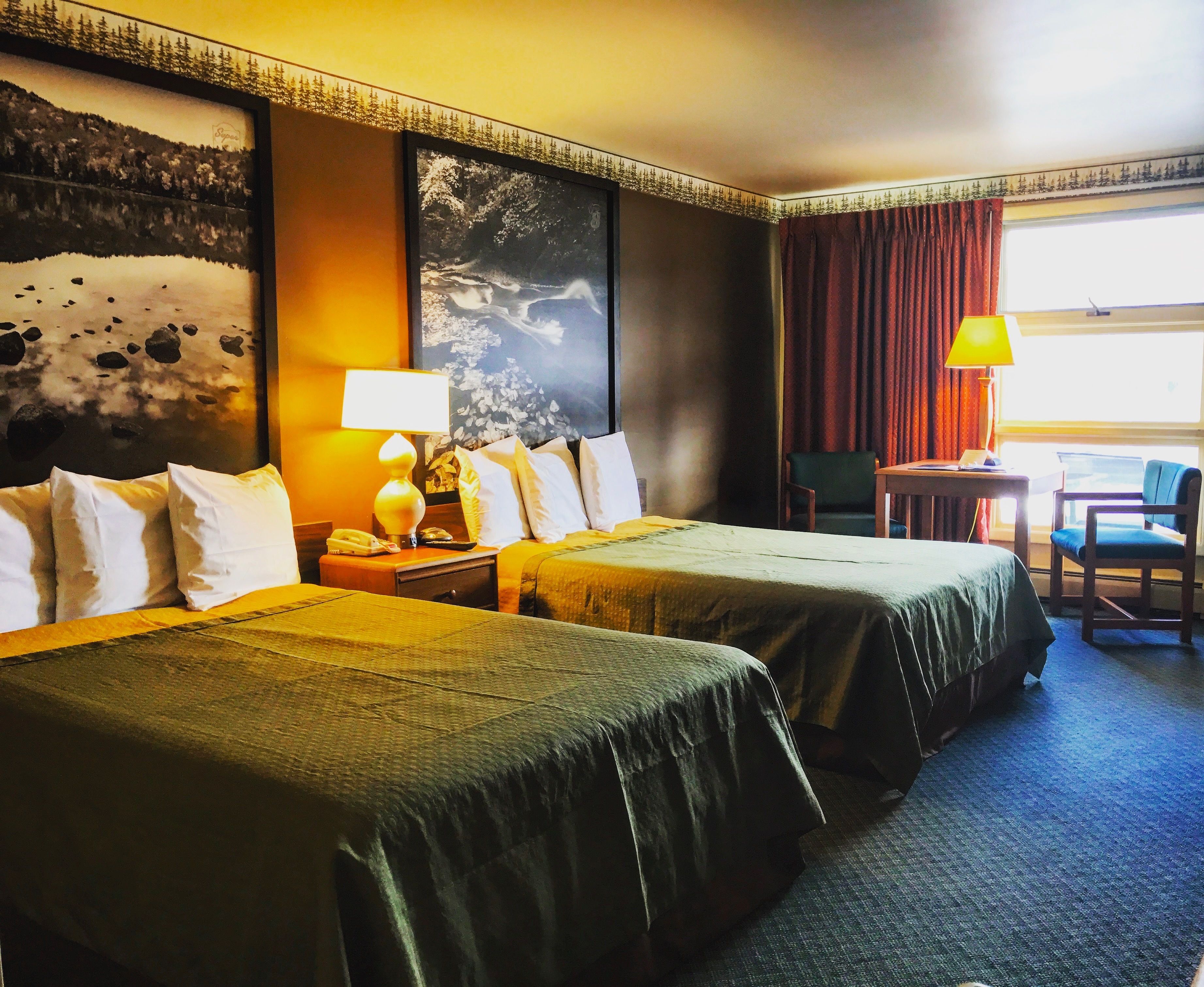A hotel room with two beds, a desk, a chair, a lamp, and a large window with curtains.