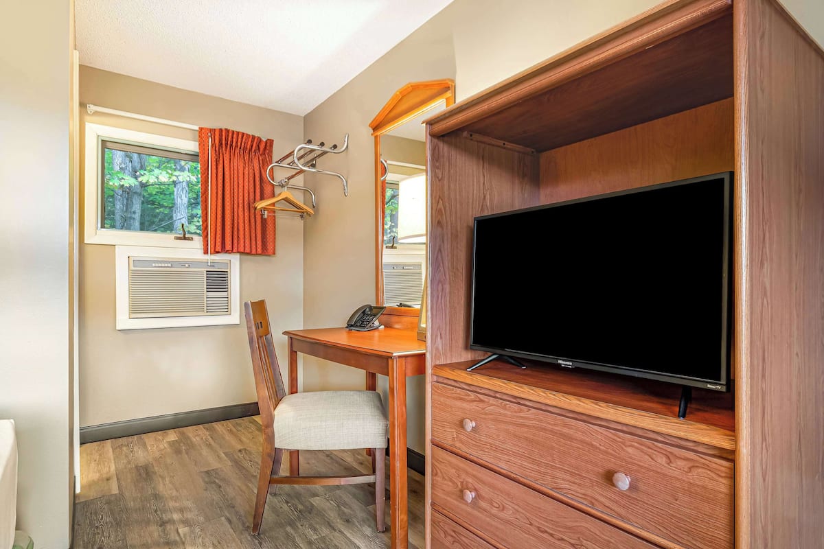 Une pièce de taille modeste, avec un bureau en bois, une chaise, un téléviseur posé sur une commode en bois, une fenêtre avec des rideaux et un radiateur fixé au mur.