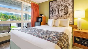 A hotel room with a large bed, a nightstand with a lamp, a coffee maker, and a view of the outdoors.