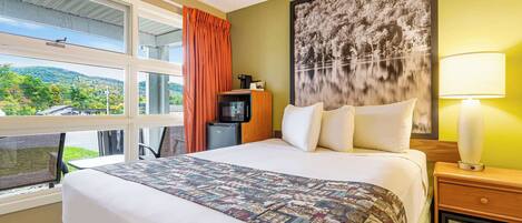 A hotel room with a large bed, a nightstand with a lamp, a coffee maker, and a view of the outdoors.