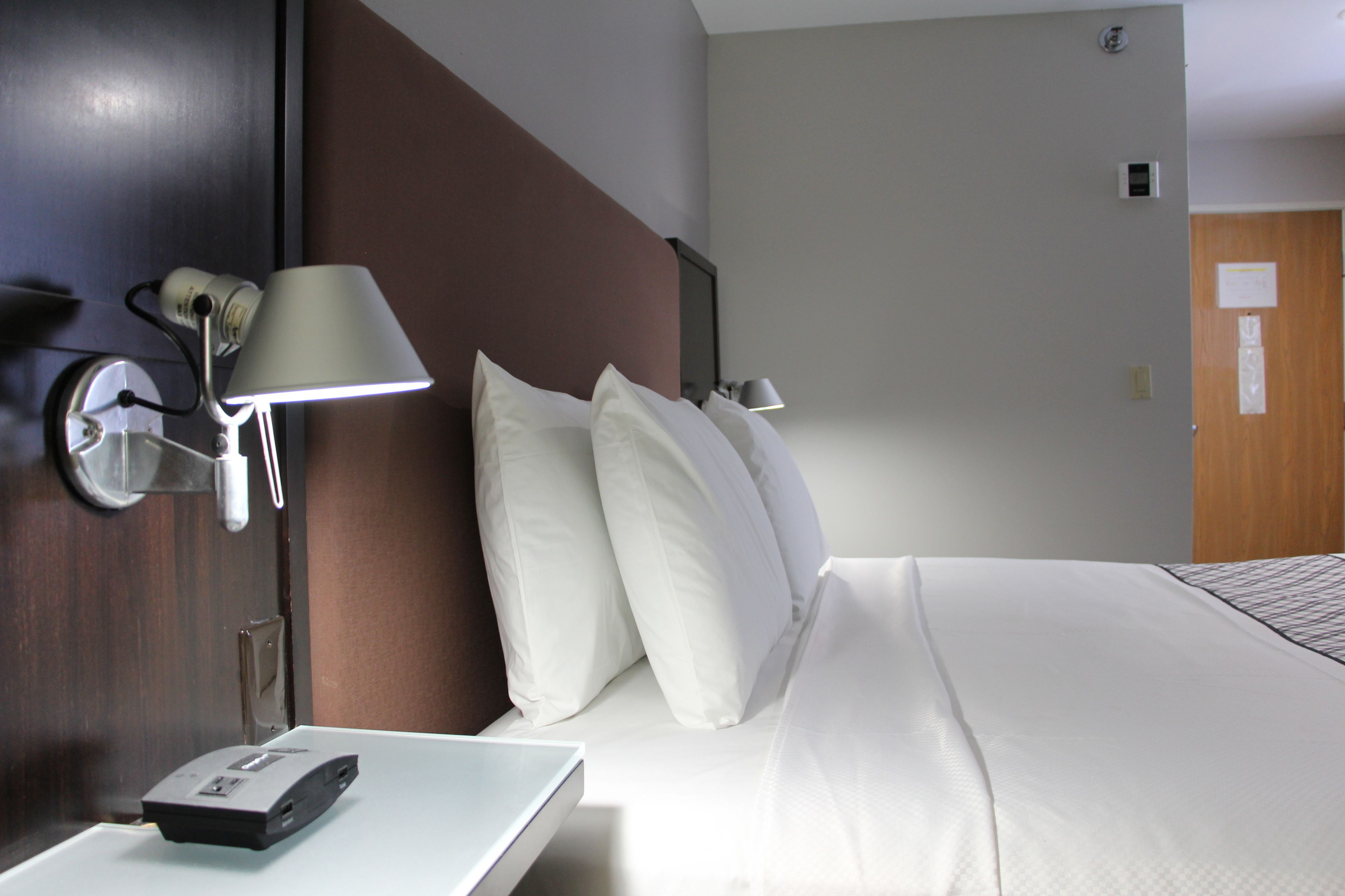 A hotel room with a bed, white bedding, a nightstand with a lamp, and a remote control.