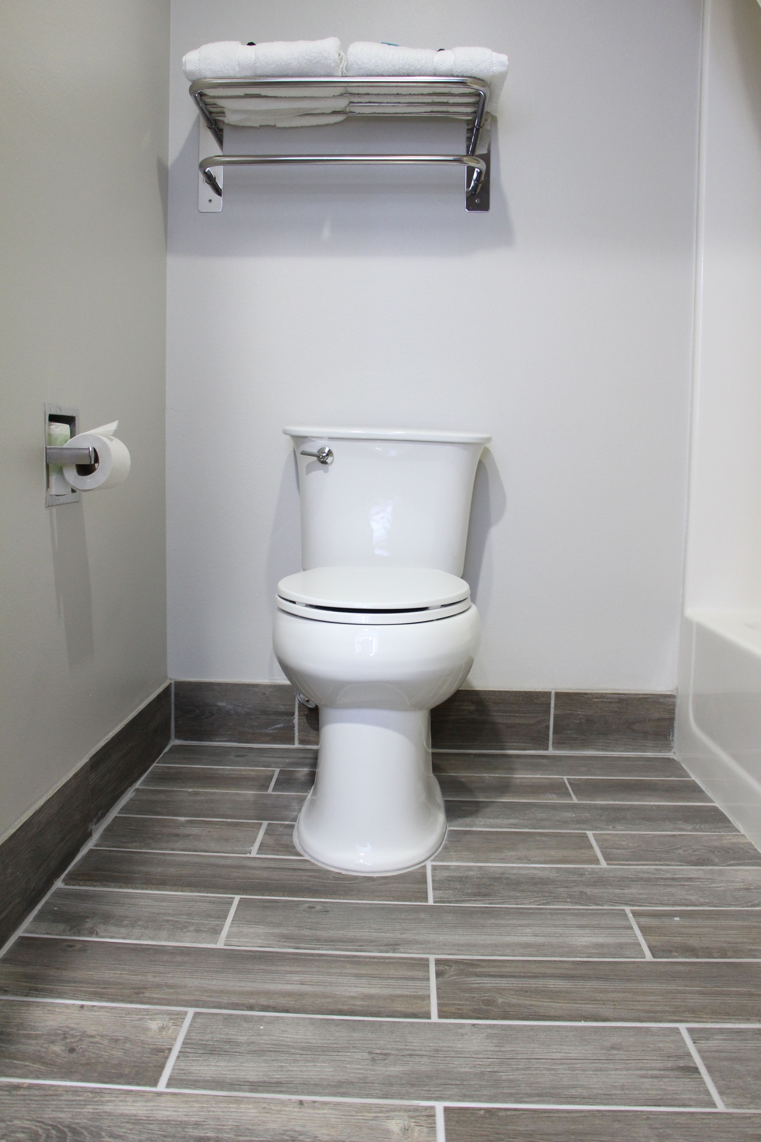 A white toilet with a metal shelf above it, grey wooden floor tiles, and a white wall.