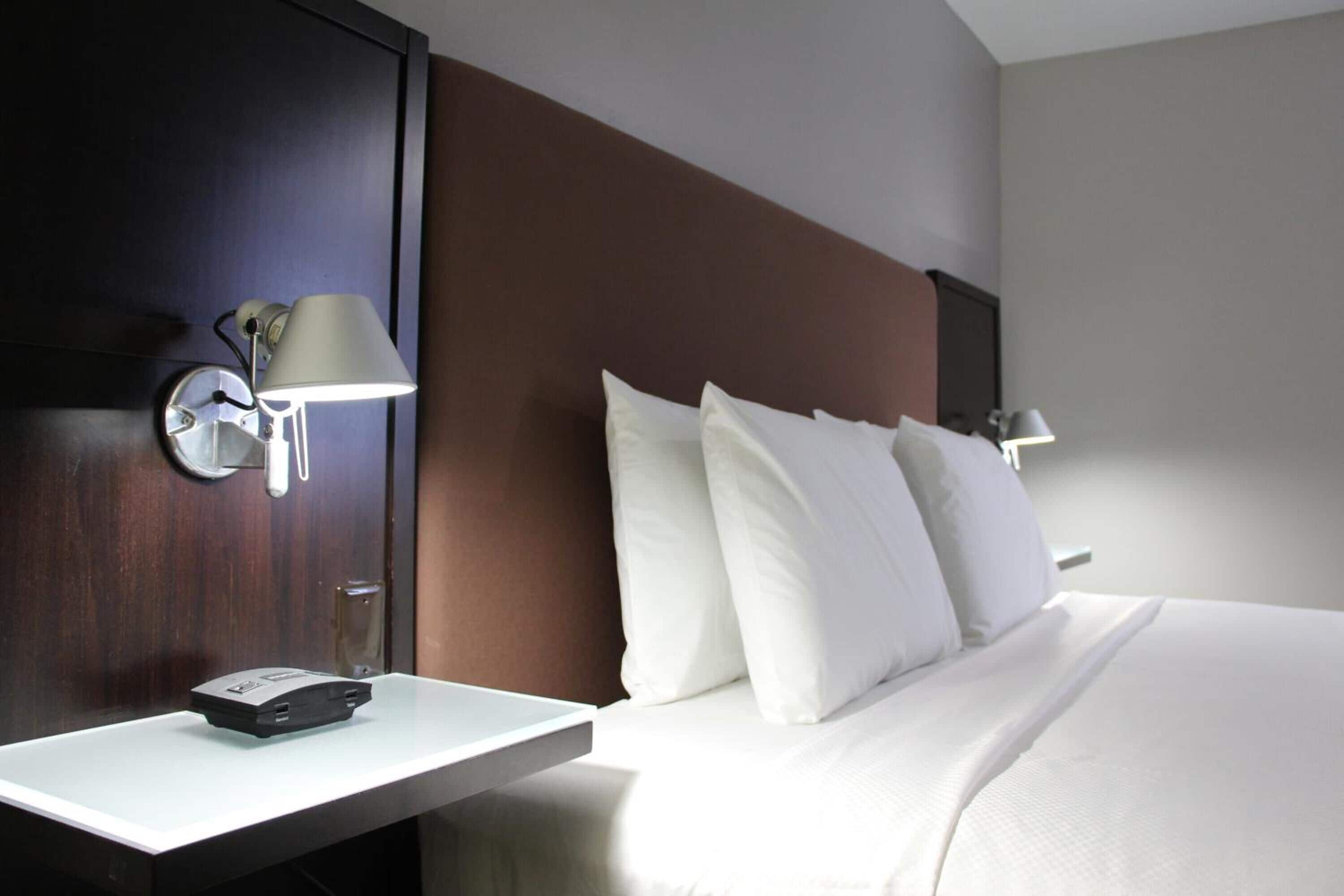 A hotel room with a bed, white bedding, a nightstand with a lamp, and a wall-mounted phone.