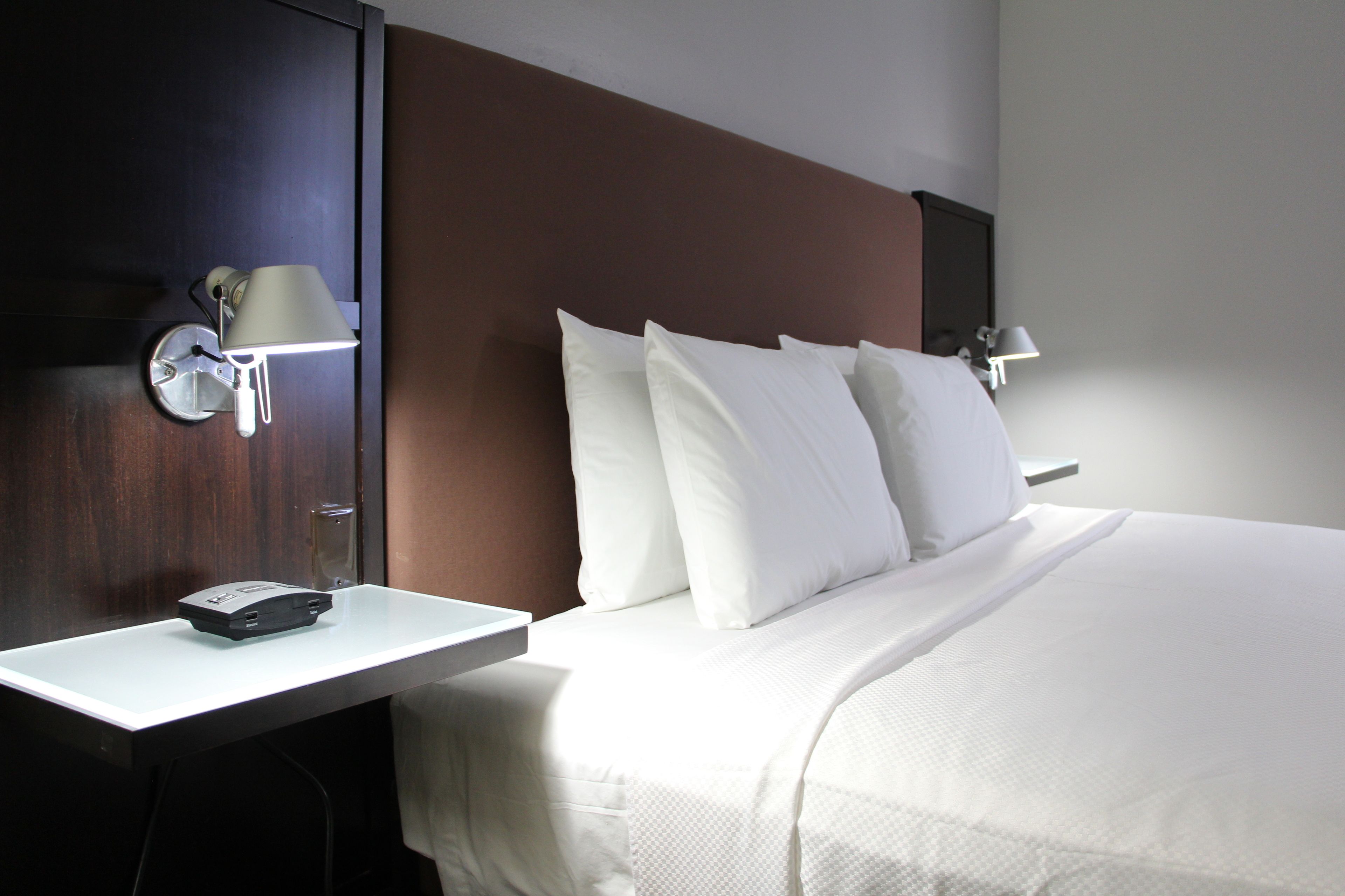 Quarto de hotel com cama, almofadas brancas, mesa de cabeceira com abajur e luminária na parede.