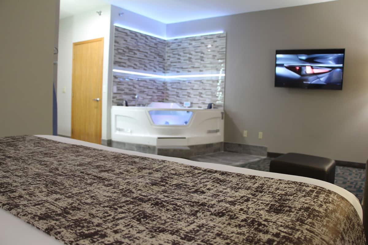 A modern hotel room with a large bed, a built-in jacuzzi, a flat-screen TV, and a stone-textured wall.