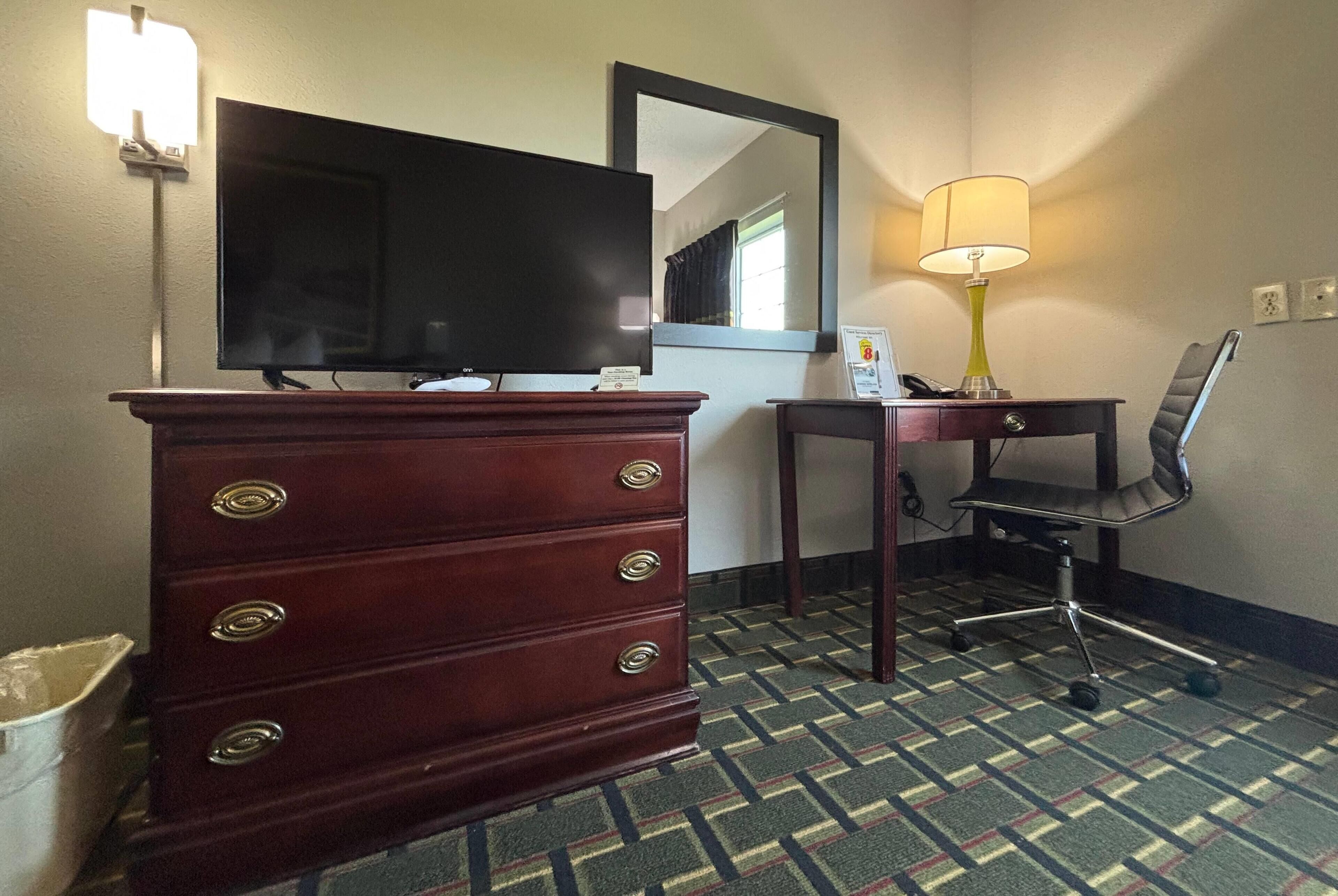A hotel room with a wooden dresser, a television, a desk with a chair, and a patterned carpet.