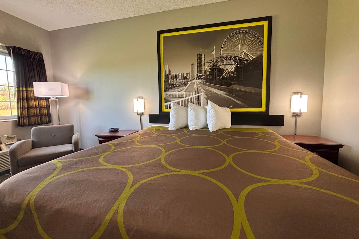 A hotel room with a large bed, two bedside lamps, a chair, a desk, and a large framed picture of a cityscape with a Ferris wheel.