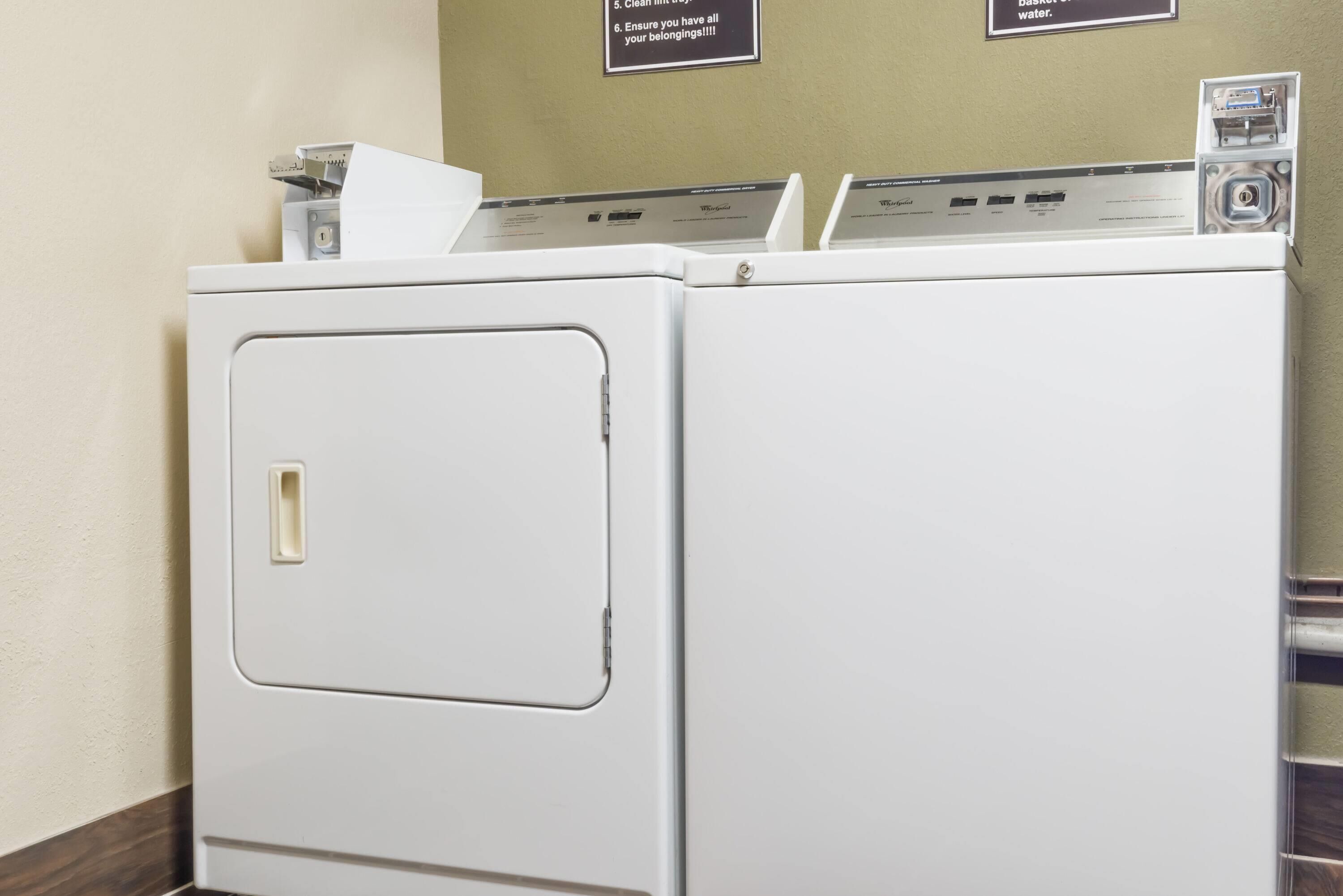 A row of white front-loading washers with control panels, set against a beige wall with two informational signs.