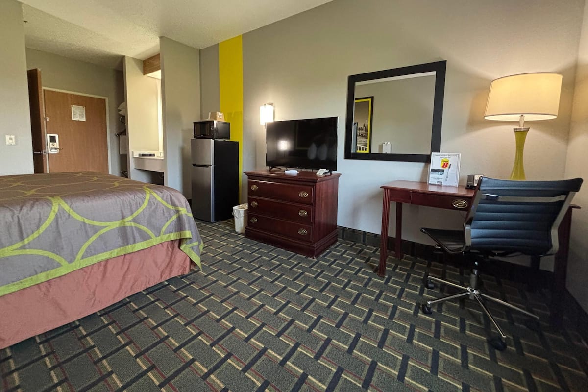 A hotel room with a bed, a desk with a chair, a television, a mirror, a lamp, and a refrigerator.