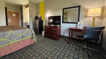 A hotel room with a bed, a desk with a chair, a television, a mirror, a lamp, and a refrigerator.