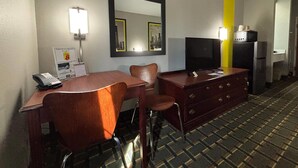 A hotel room with a wooden desk, two chairs, a mirror, a television, and a dresser.