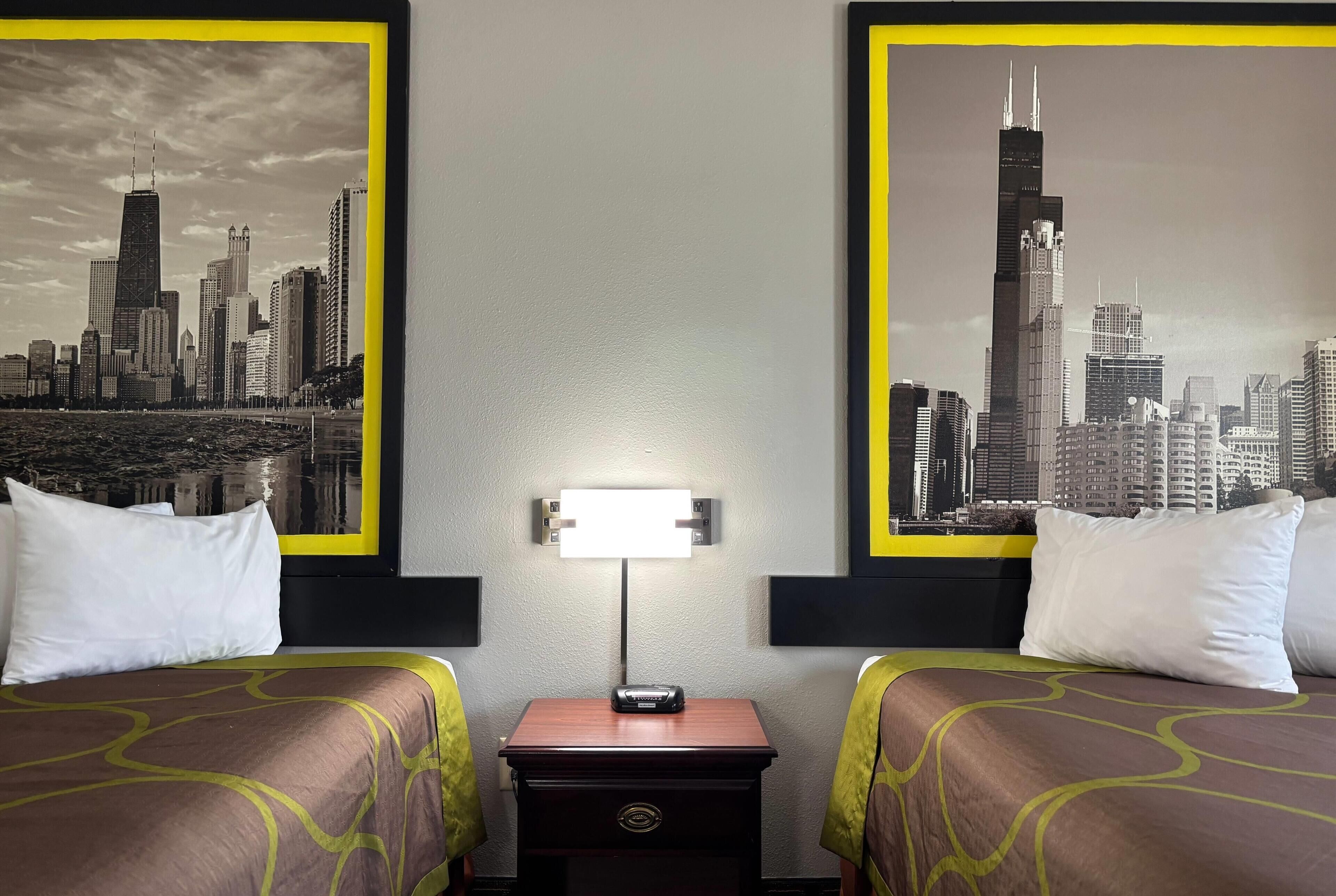 A hotel room with two beds, a nightstand, a lamp, and two large cityscape posters on the walls.