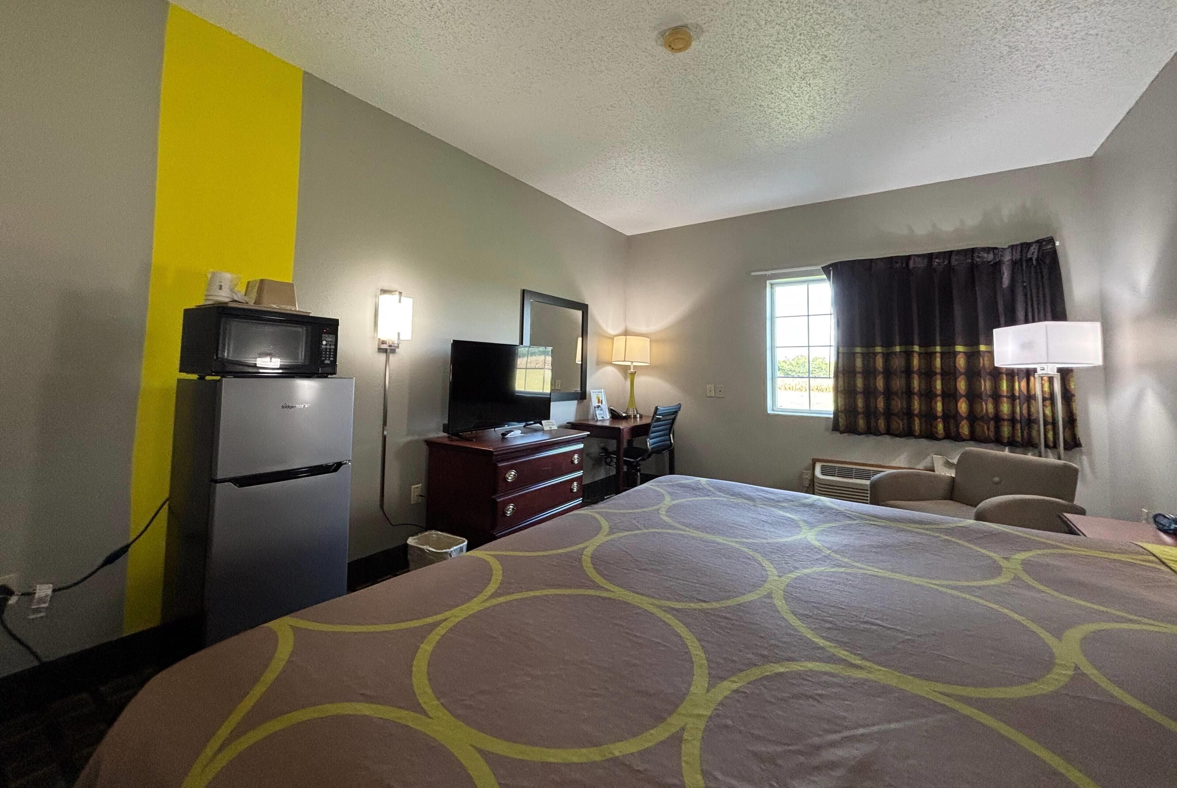 Habitación de hotel con cama, escritorio, televisor, refrigerador y microondas, y una ventana con cortinas.