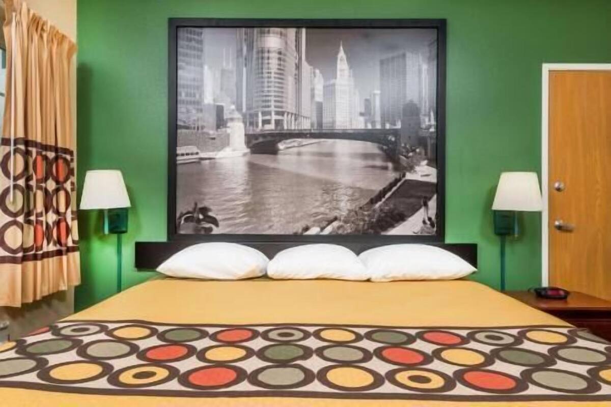 A hotel room with a bed, a large framed cityscape picture on the wall, two bedside lamps, and a patterned bedspread.