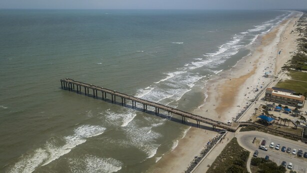 Une vue aérienne d’une plage avec une longue jetée qui s’avance dans l’océan, bordée d’une plage de sable où se trouvent des bâtiments et des voitures garées.