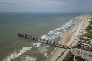 Vista aérea de una playa con un muelle largo que se adentra en el océano, junto a una orilla arenosa con edificios y coches estacionados.