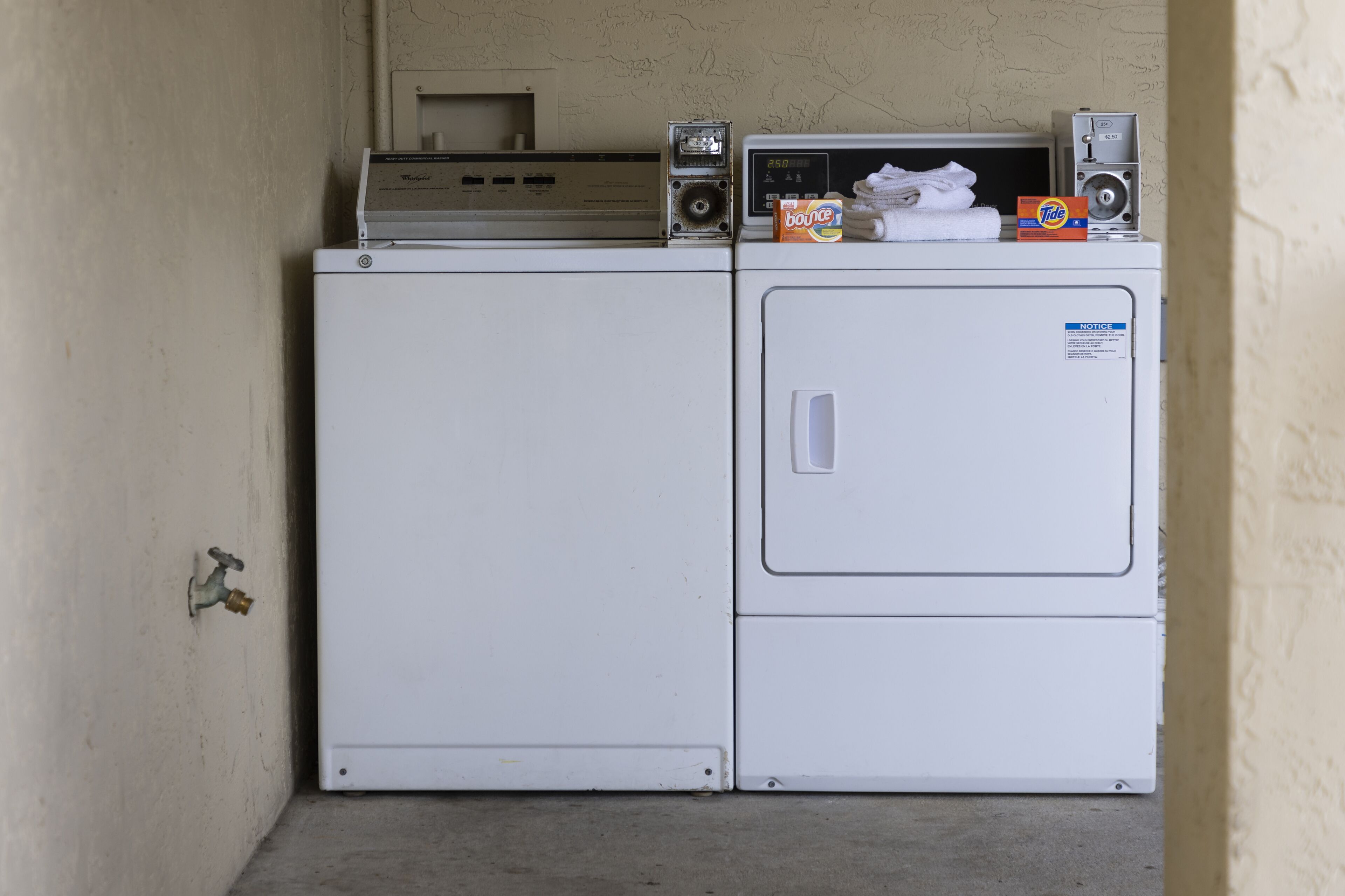 A compact laundry area with a front-loading washer and dryer, a small shelf with a speaker and a box of Tide detergent, and a visible water tap.