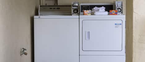 A compact laundry area with a front-loading washer and dryer, a small shelf with a speaker and a box of Tide detergent, and a visible water tap.
