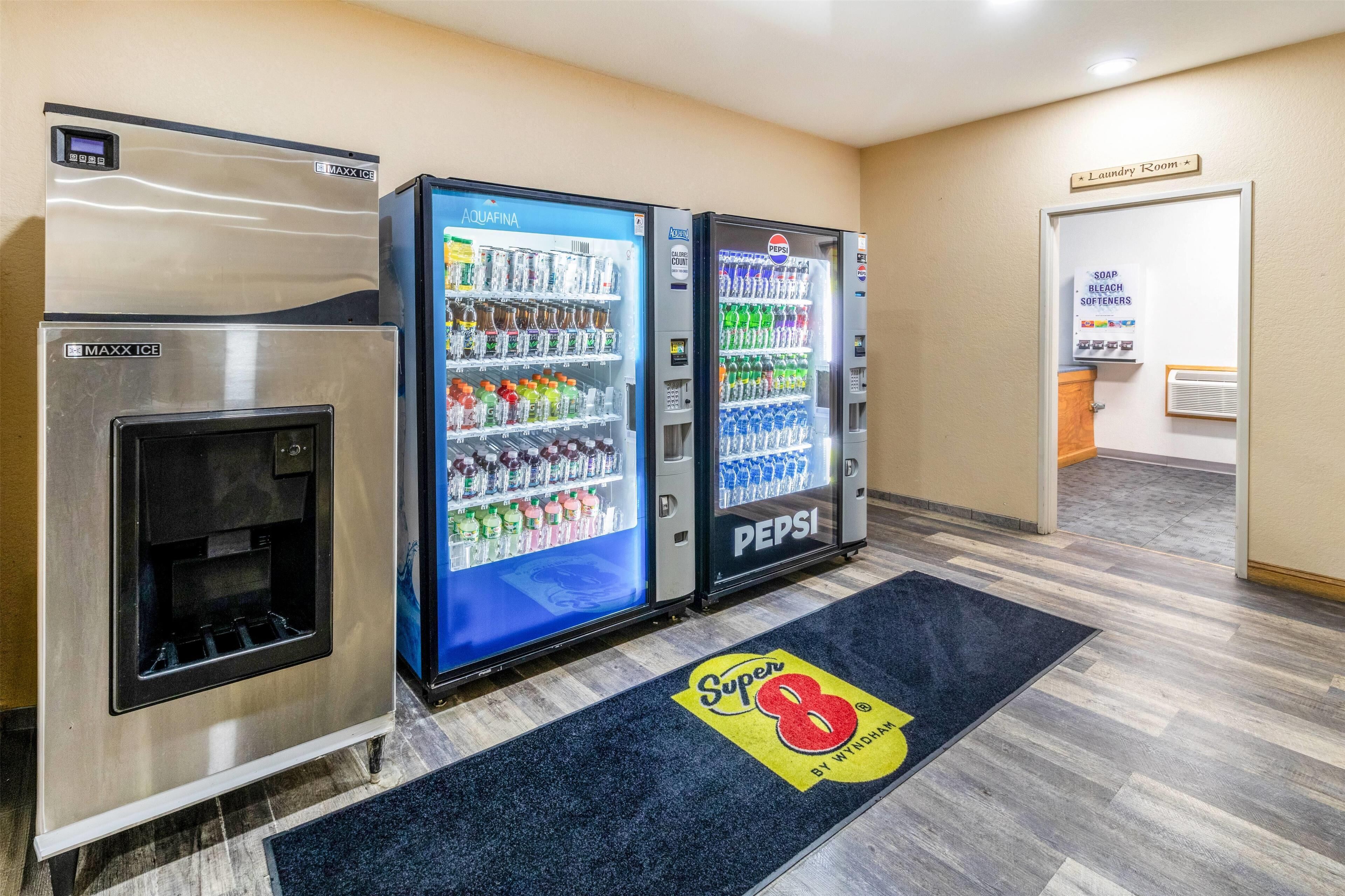 Uma área de autoatendimento com máquinas de gelo MAXX ICE, AQUAFINA e PEPSI, o logotipo Super 8 by Wyndham no tapete e uma placa indicando a lavanderia.