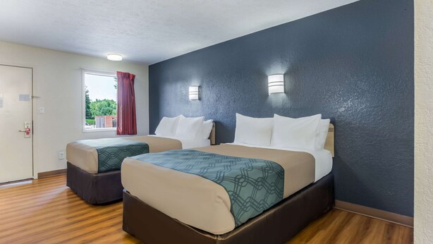 Habitación de hotel con dos camas, piso de madera, ventanal con cortinas rojas e iluminación en la pared.