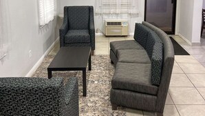 Sala de estar con sofá, sillón y mesa de centro. Se ve una ventana con cortinas blancas, un radiador y una puerta que da a otra habitación.