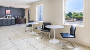 Una cafetería moderna con zona de cocina, tres mesas redondas con sillas y grandes ventanales con vistas al exterior.