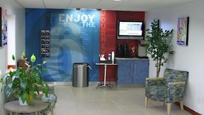 A reception area with a blue wall, a television, a trash bin, a small table with a plant, and a patterned chair. - Motel 6 Urbana, IL (Urbana)