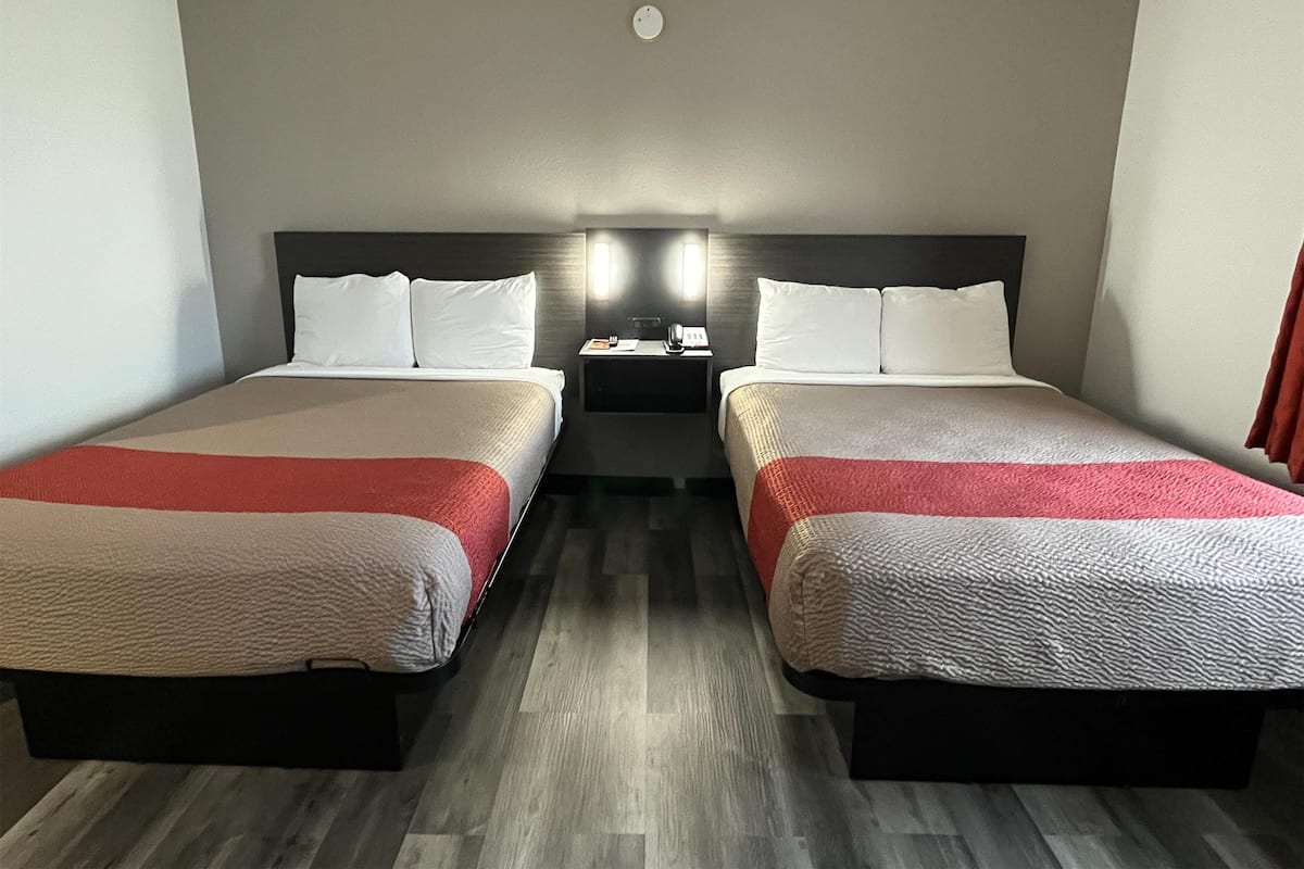 A hotel room with two beds, each with a red and white bedspread, a nightstand with a lamp, and a wall-mounted light fixture.