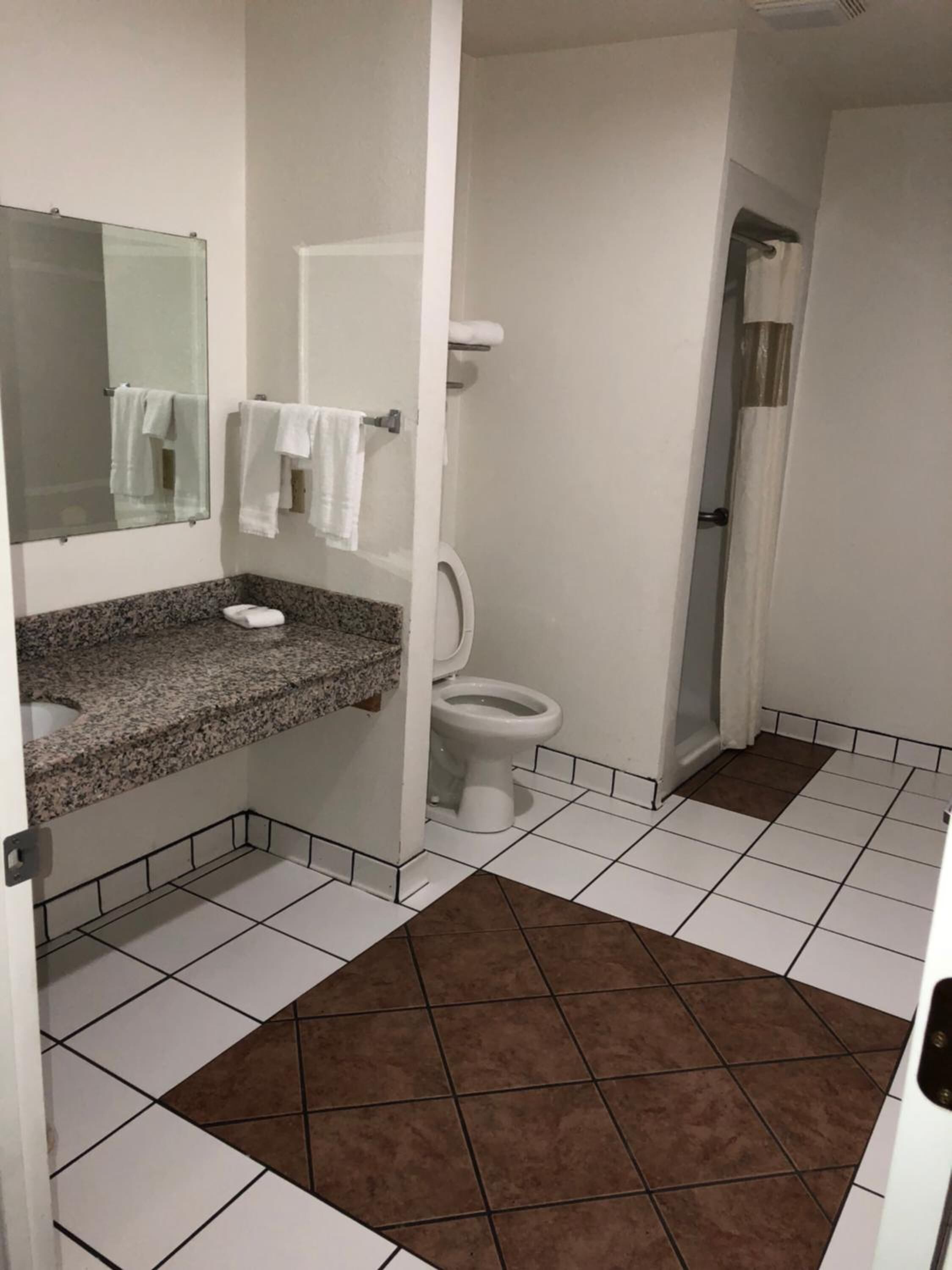 A bathroom with a granite countertop, white tiles, a toilet, a shower, and a mirror.