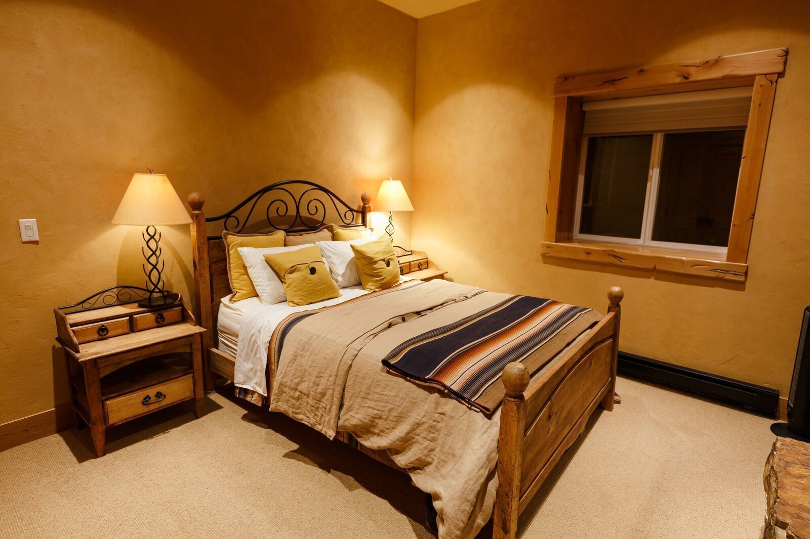 A bedroom with a wooden bed, two bedside tables with lamps, a window, and a carpeted floor.