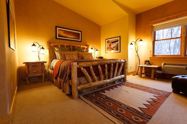 A cozy bedroom with a rustic wooden bed, a small bedside table, a bench, and a large window with a view of trees.