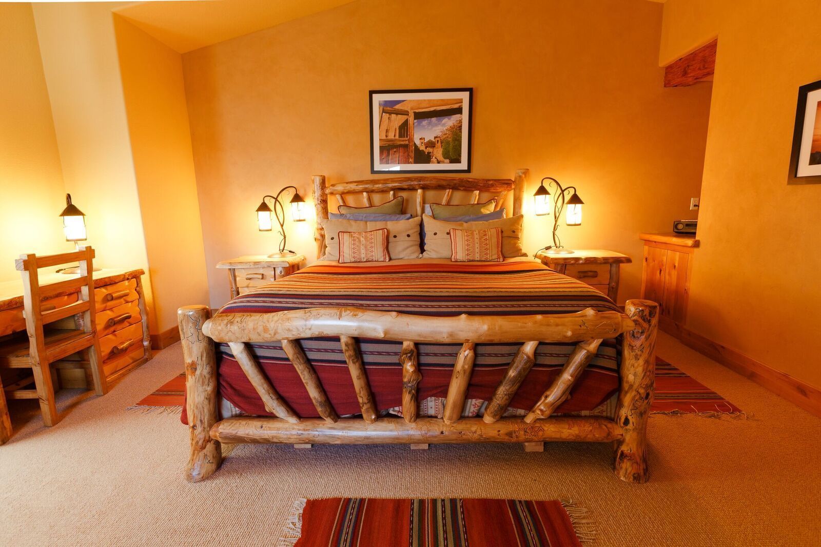 A rustic bedroom with a wooden bed frame, a large bedspread, two bedside lamps, a wooden nightstand, a wooden dresser, and a framed picture on the wall.