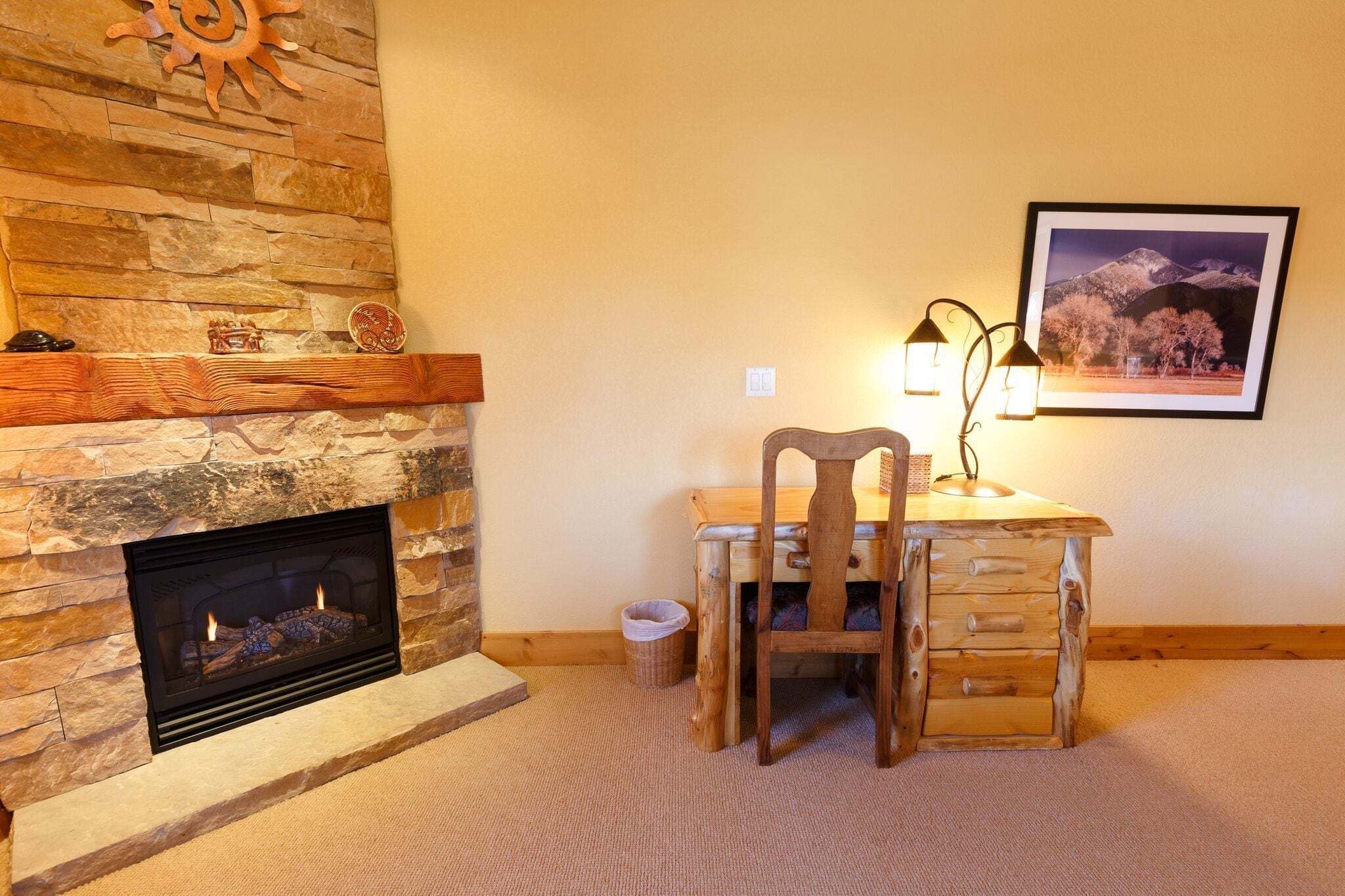 A cozy room with a stone fireplace, a wooden desk with a chair, a framed picture on the wall, and a small trash can.