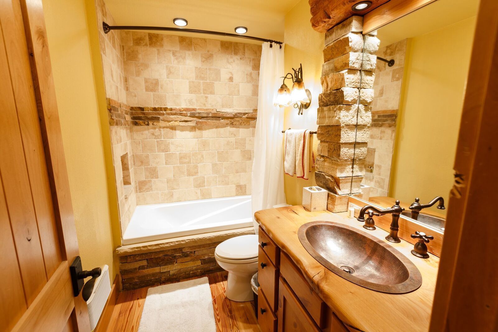Un baño con ducha de azulejos de piedra, tocador de madera con un lavamanos grande y un espejo con un detalle de pared de piedra.