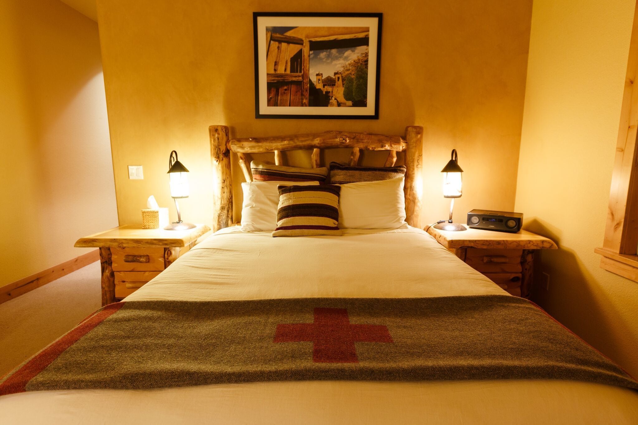 A hotel room with a wooden headboard, a bed with a white comforter and a red cross blanket, two bedside tables with lamps, and a framed picture on the wall.