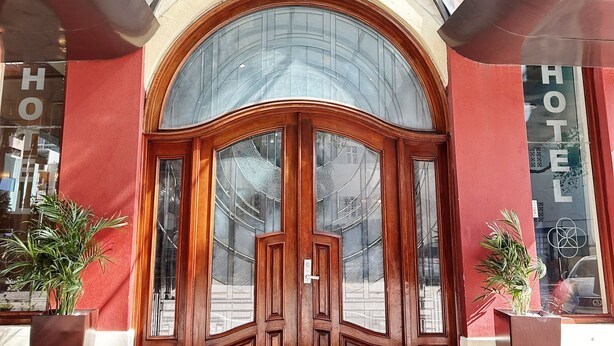 Entrada de hotel con marquesina de vidrio, puertas de madera y mostrador de recepción.