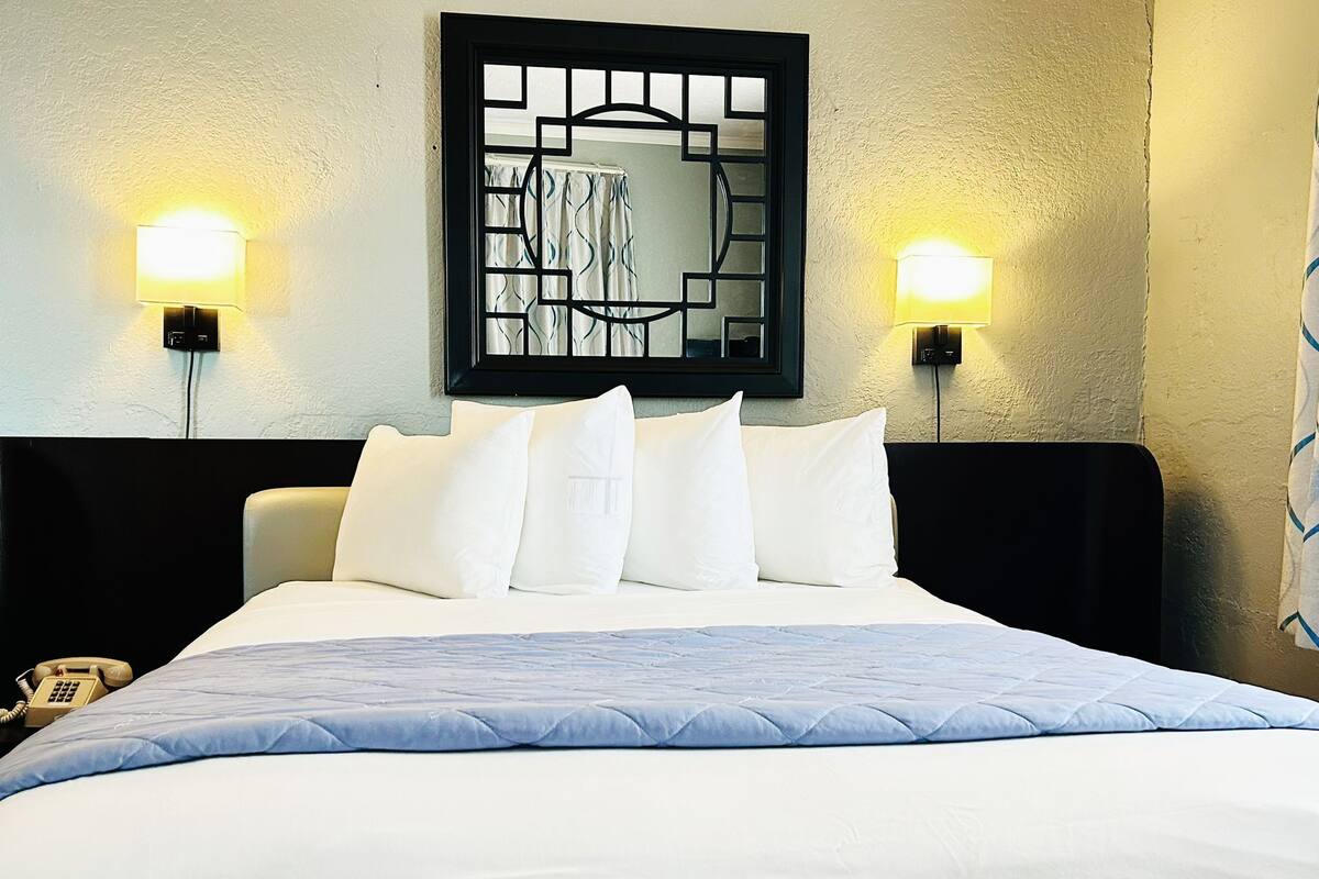 Een hotelkamer met een bed, witte kussens, een hoofdbord, twee wandlampen en een telefoon op het nachtkastje.