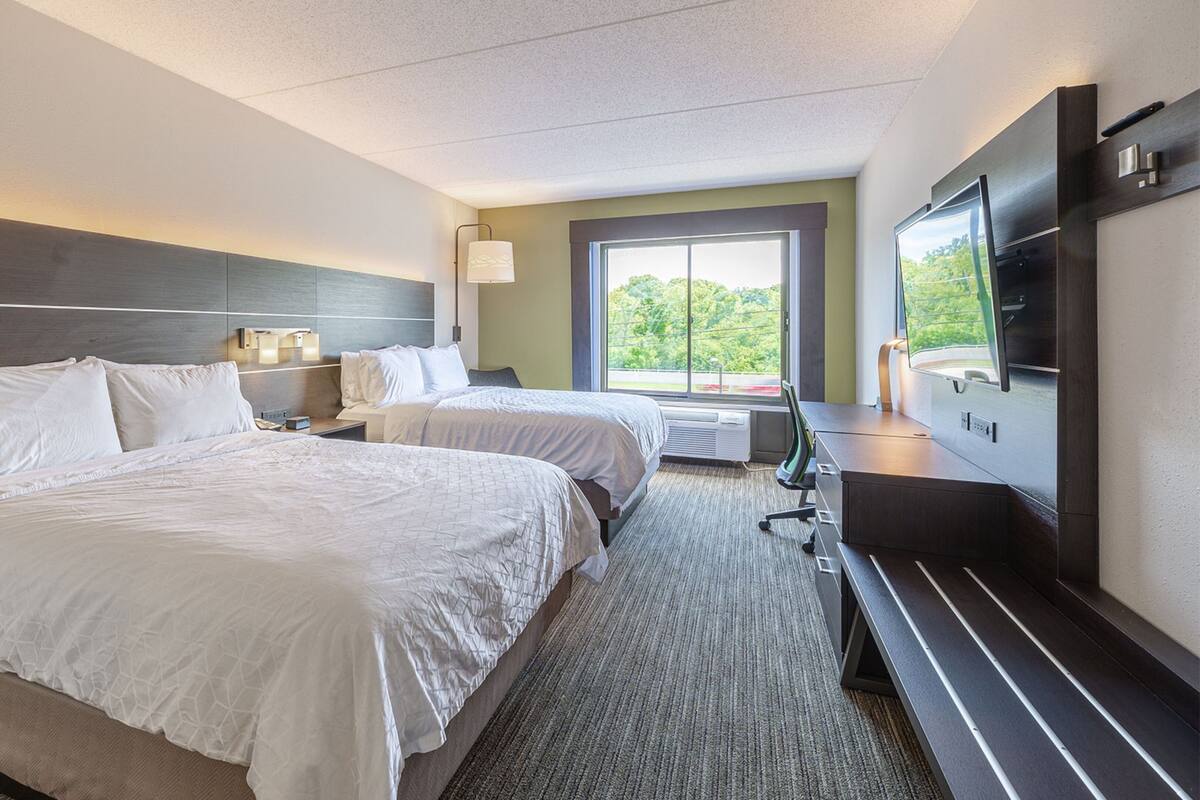 A hotel room with two beds, a desk, a TV, and a large window with a view of greenery.