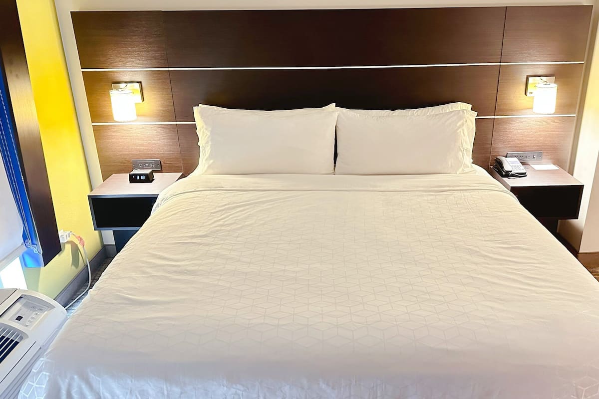 A hotel room with a large bed, two bedside tables with lamps, a wall-mounted air conditioner, and a window with a curtain.