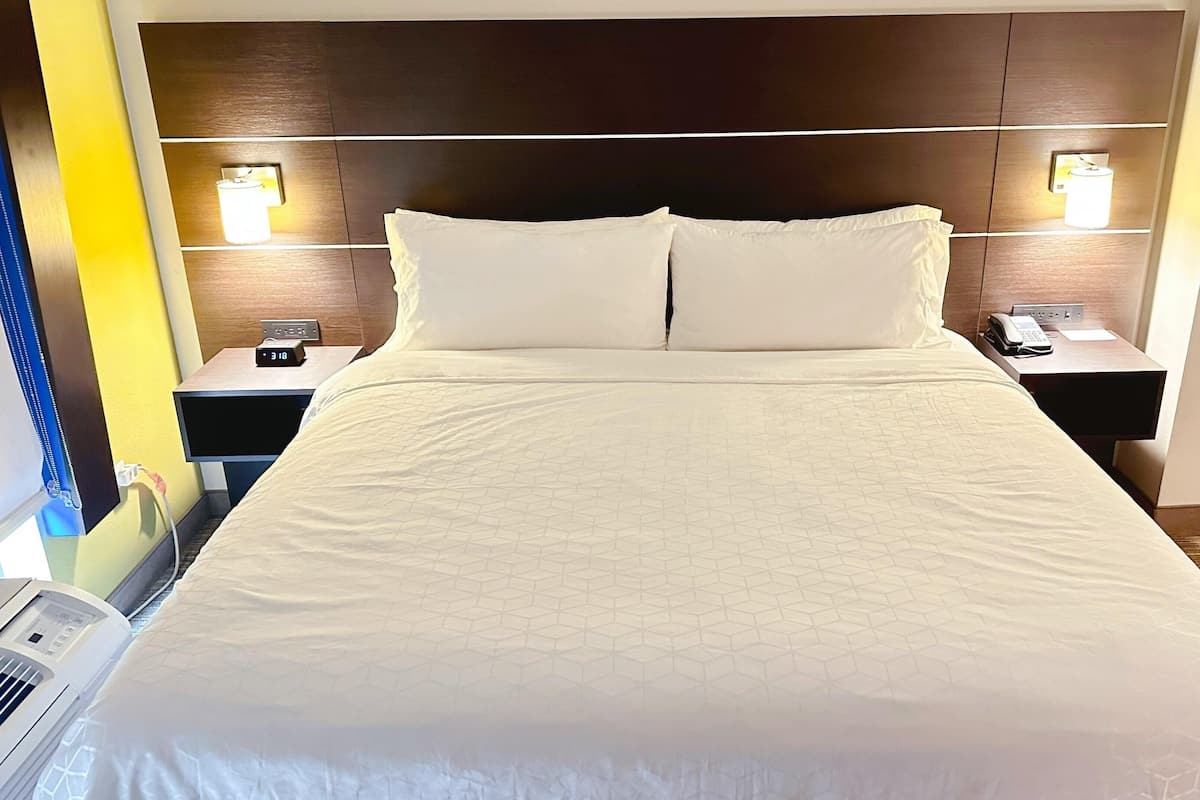 A hotel room with a large bed, two bedside tables with lamps, a wall-mounted air conditioner, and a bedside phone.